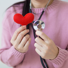 Woman wearing a pink sweater holding a stethoscope up next to a small red felt heart